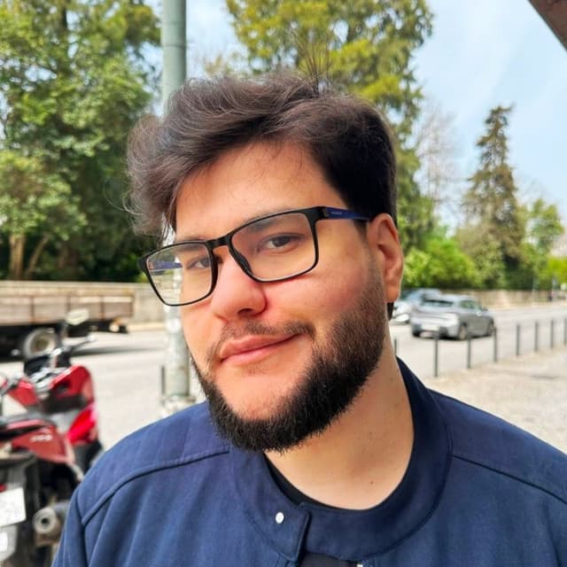 Me, a man with short dark hair, glasses, and a trimmed beard, standing outdoors near a street with parked motorcycles and cars, wearing a navy jacket and smiling slightly at the camera. Trees and a sidewalk are visible in the background.